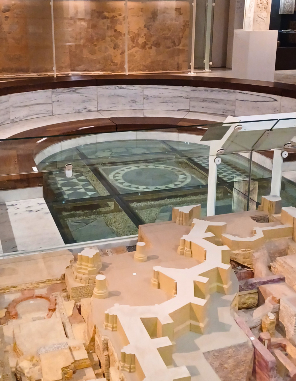 Ausstellungsbereich mit einer großen Bodenvitrine, die einen antiken Mosaikboden unter Glas zeigt. Darum herum verläuft ein halbrunder Sitz- oder Präsentationsbereich aus Stein und Holz. Im Vordergrund befindet sich ein archäologisches Modell mit rekonstruierten Gebäudestrukturen und Mauern.