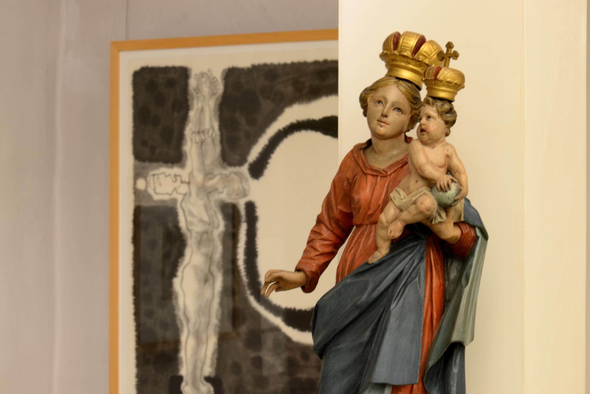 Blick in die Ausstellung 'Menschenskinder' mit einer Maria Immaculata im Vordergrund, dahinter eine Tuschzeichnung von Klaus Maßem zu einem Hexenprozeß.entius sowie mit einer Josefsstatue mit Jesuskind.