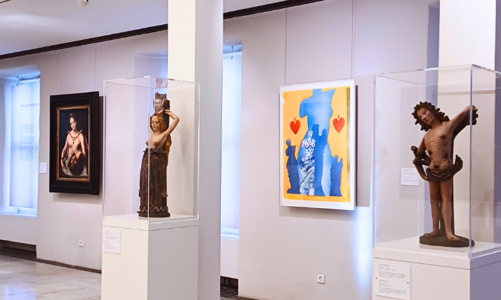 Heller Ausstellungsraum in einem Museum mit Skulpturen und Gemälden: Zwei historische Holzfiguren in Glasvitrinen stehen auf weißen Sockeln, daneben hängen ein klassisches Gemälde und ein modernes farbenfrohes Kunstwerk mit Blau‑Gelb‑Kontrasten und roten Herzen. Der Museumsraum ist großzügig beleuchtet und zeigt eine vielfältige Kunstsammlung.