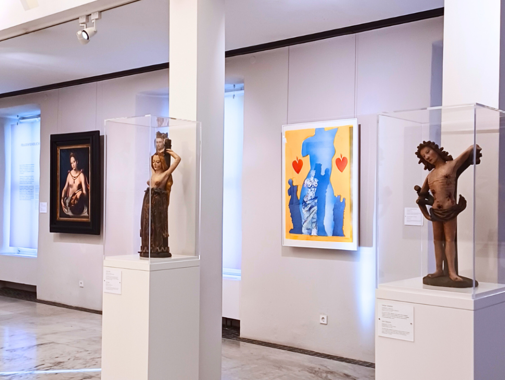 Heller Ausstellungsraum in einem Museum mit Skulpturen und Gemälden: Zwei historische Holzfiguren in Glasvitrinen stehen auf weißen Sockeln, daneben hängen ein klassisches Gemälde und ein modernes farbenfrohes Kunstwerk mit Blau‑Gelb‑Kontrasten und roten Herzen. Der Museumsraum ist großzügig beleuchtet und zeigt eine vielfältige Kunstsammlung.
