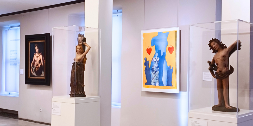 Heller Ausstellungsraum in einem Museum mit Skulpturen und Gemälden: Zwei historische Holzfiguren in Glasvitrinen stehen auf weißen Sockeln, daneben hängen ein klassisches Gemälde und ein modernes farbenfrohes Kunstwerk mit Blau‑Gelb‑Kontrasten und roten Herzen. Der Museumsraum ist großzügig beleuchtet und zeigt eine vielfältige Kunstsammlung.