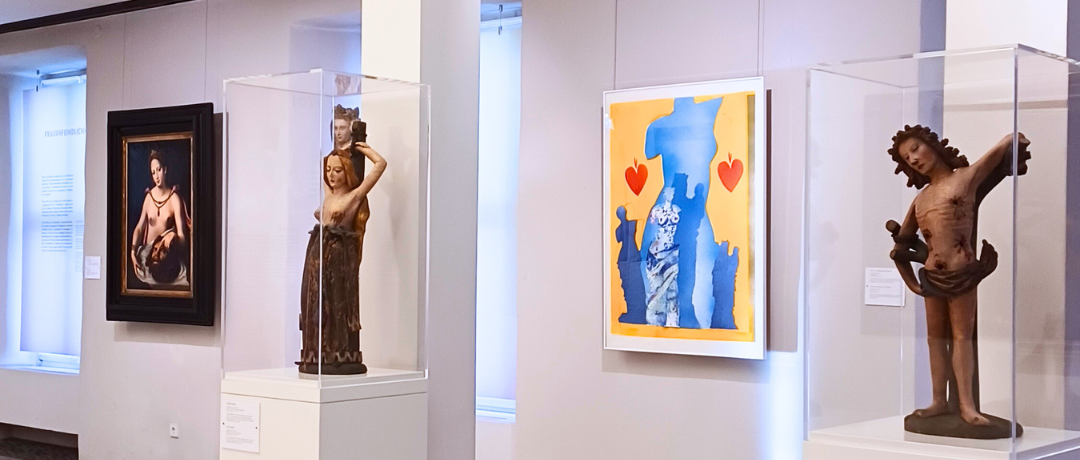 Heller Ausstellungsraum in einem Museum mit Skulpturen und Gemälden: Zwei historische Holzfiguren in Glasvitrinen stehen auf weißen Sockeln, daneben hängen ein klassisches Gemälde und ein modernes farbenfrohes Kunstwerk mit Blau‑Gelb‑Kontrasten und roten Herzen. Der Museumsraum ist großzügig beleuchtet und zeigt eine vielfältige Kunstsammlung.