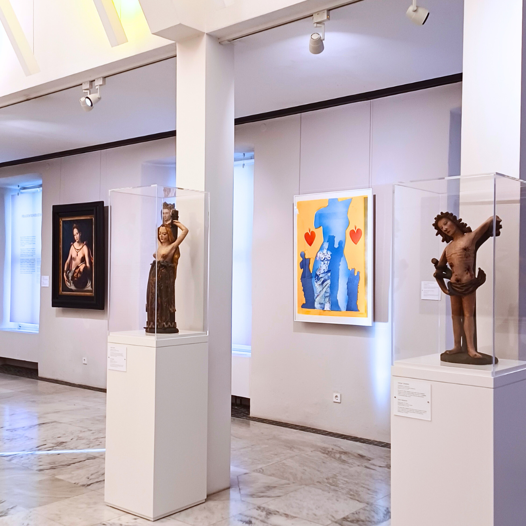 Heller Ausstellungsraum in einem Museum mit Skulpturen und Gemälden: Zwei historische Holzfiguren in Glasvitrinen stehen auf weißen Sockeln, daneben hängen ein klassisches Gemälde und ein modernes farbenfrohes Kunstwerk mit Blau‑Gelb‑Kontrasten und roten Herzen. Der Museumsraum ist großzügig beleuchtet und zeigt eine vielfältige Kunstsammlung.