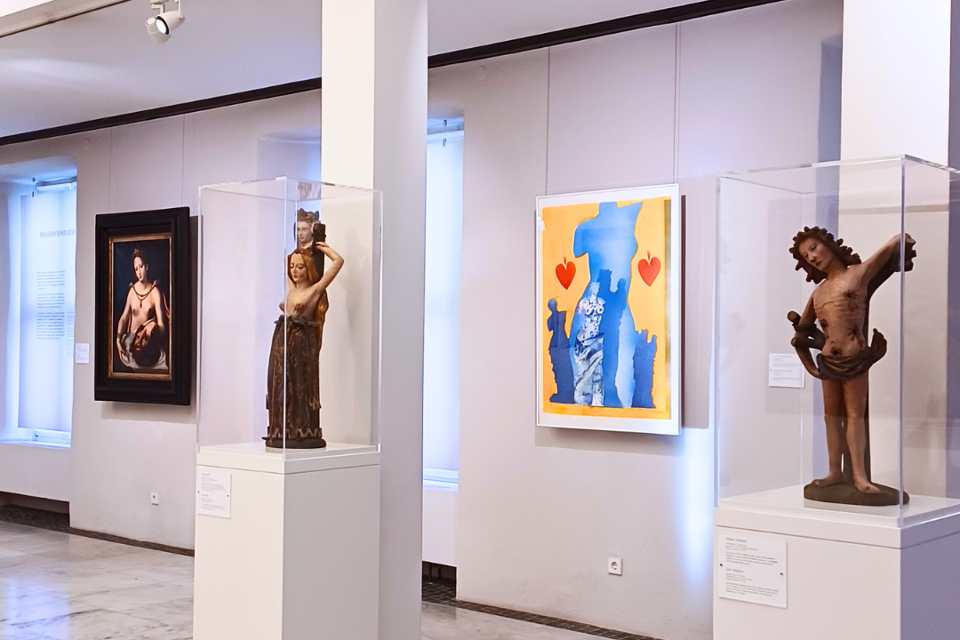 Design ohne Titel (10) (c) Museum am Dom/ Sabrina Birkel Heller Ausstellungsraum in einem Museum mit Skulpturen und Gemälden: Zwei historische Holzfiguren in Glasvitrinen stehen auf weißen Sockeln, daneben hängen ein klassisches Gemälde und ein modernes farbenfrohes Kunstwerk mit Blau‑Gelb‑Kontrasten und roten Herzen. Der Museumsraum ist großzügig beleuchtet und zeigt eine vielfältige Kunstsammlung.