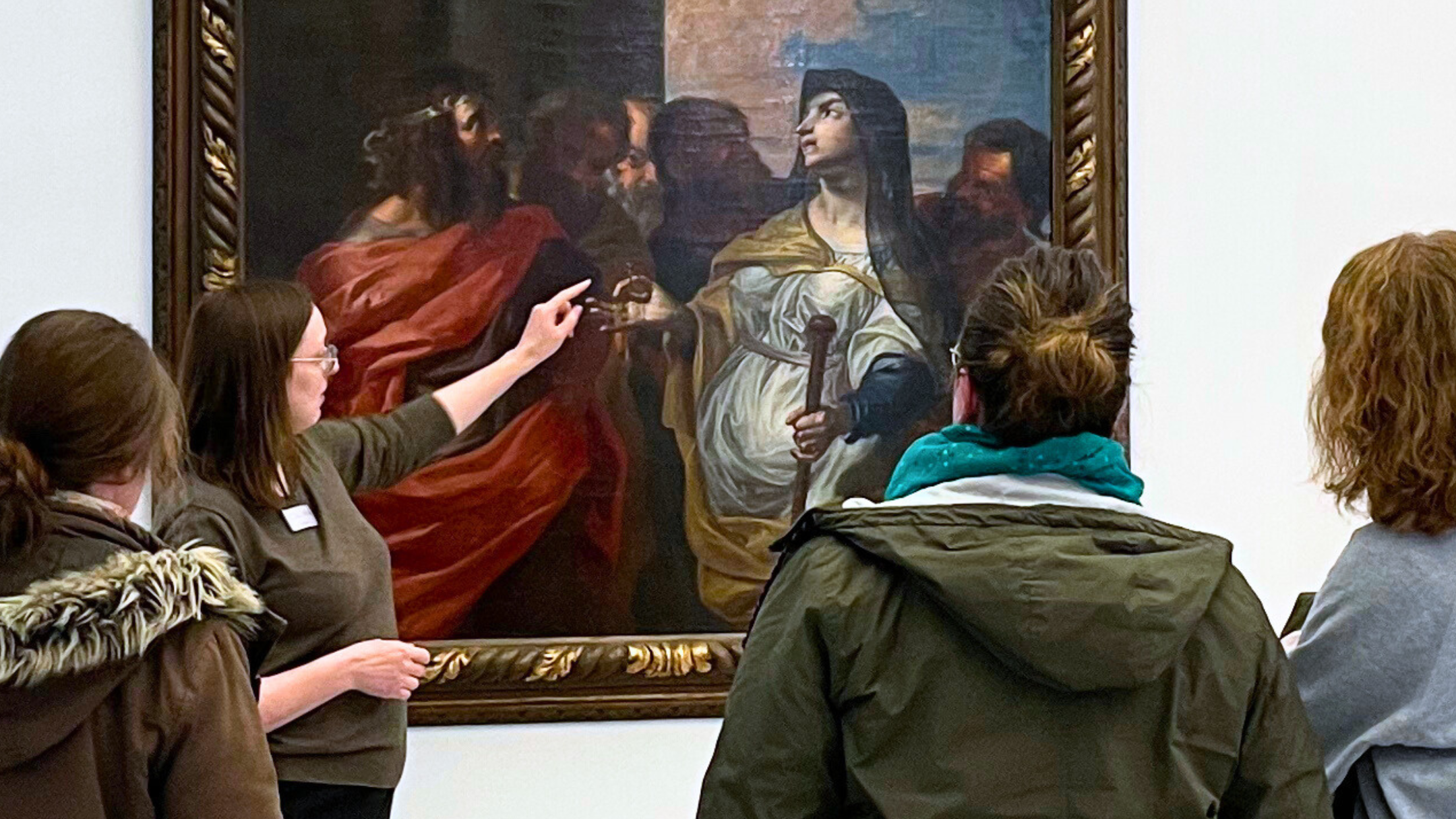 Personen bei einer Museumsführung vor einem Gemälde. Die Gruppenführerin zeigt mit ihrem Finger auf ein Detail im Bild.