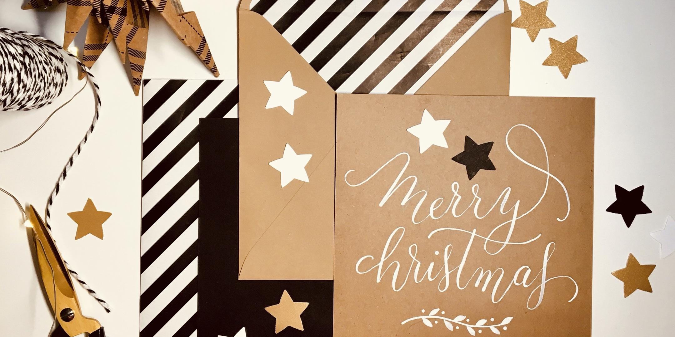 Schreib und Bastelmaterial für einen Kalligraphie Workshop. Ausgeschnittene Papier-Sterne, ein gefalteter Papier-Weihnachtsbaum, Schere und Pfaden, ein Briefumschlag und eine Karte auf der in Kalligraphie Marry Christmas steht.
