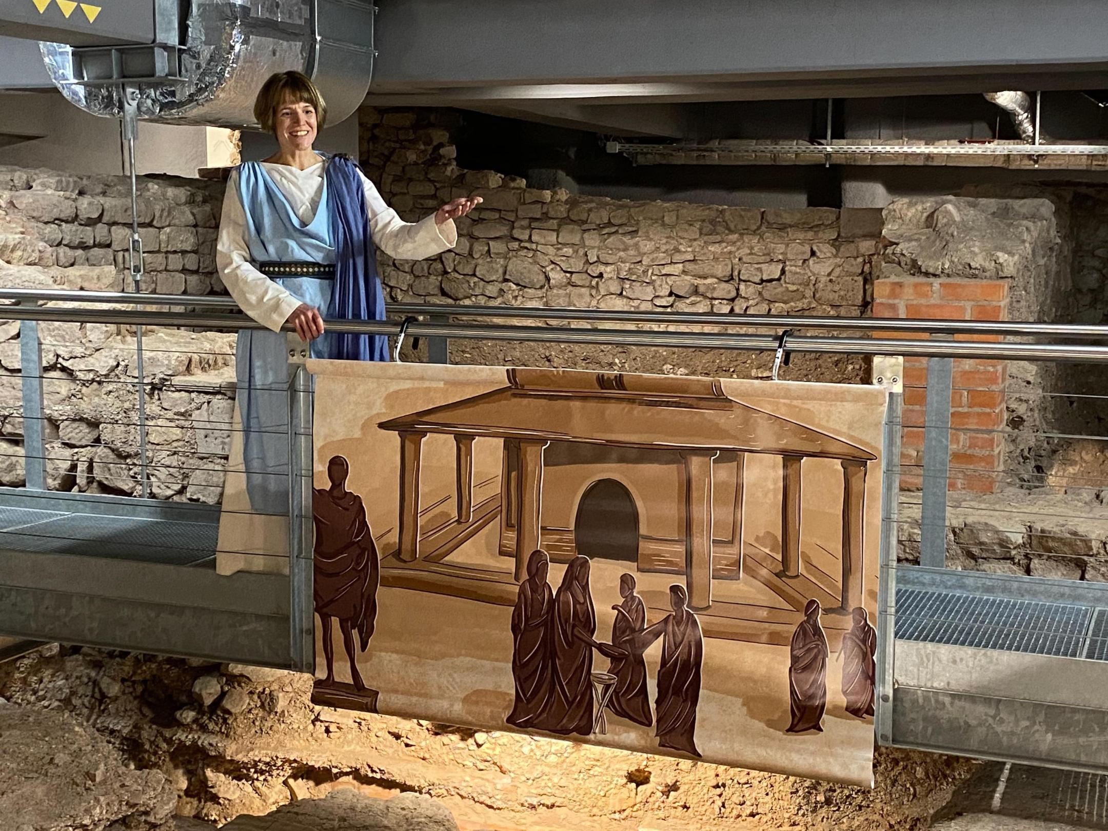 Person in historischer Gewandung steht auf einer Stegkonstruktion vor einer Mauer mit antiken Ausgrabungsresten. Am Geländer hängt eine Illustration eines antiken Gebäudes mit mehreren Menschen.