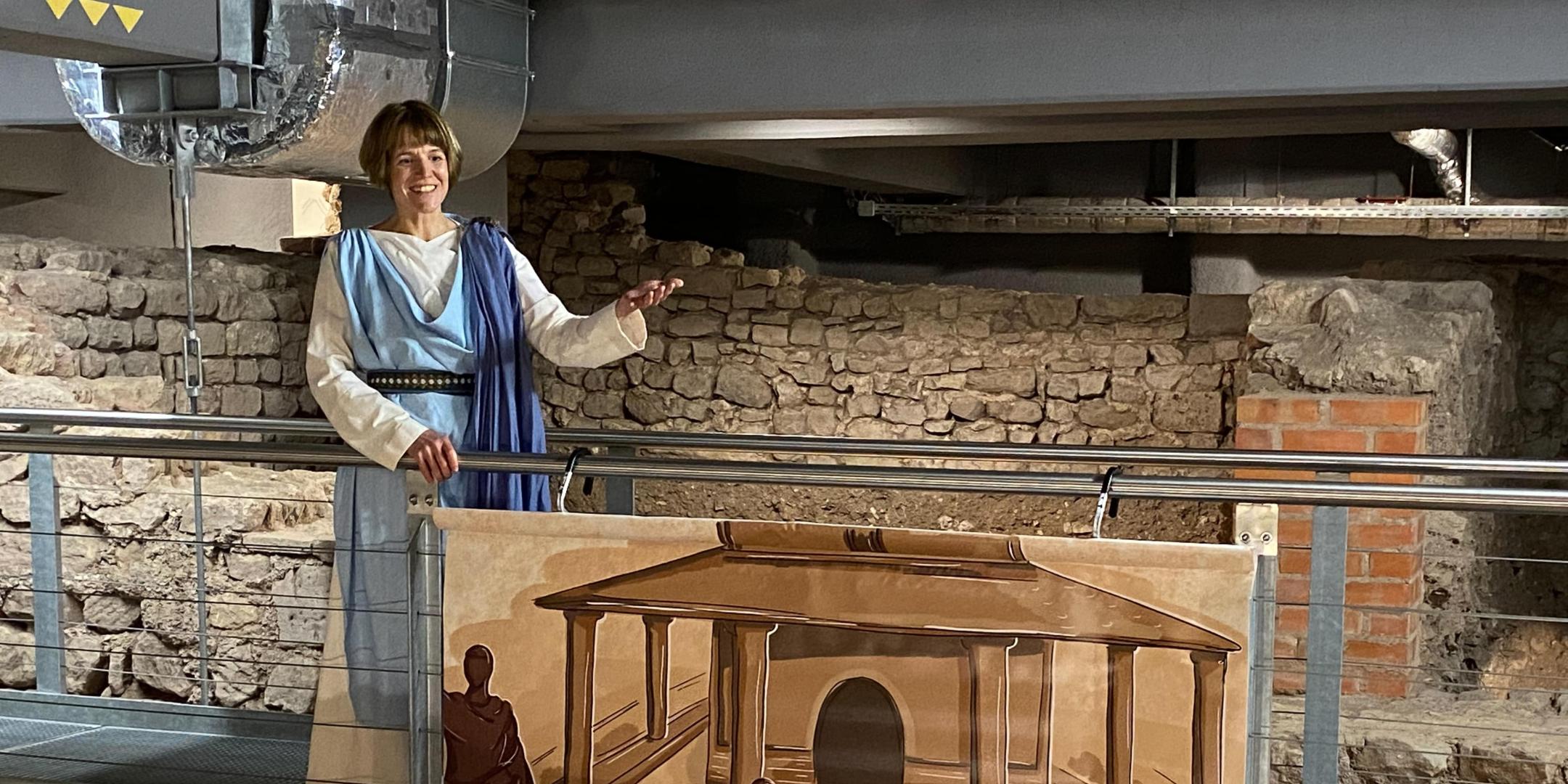 Person in historischer Gewandung steht auf einer Stegkonstruktion vor einer Mauer mit antiken Ausgrabungsresten. Am Geländer hängt eine Illustration eines antiken Gebäudes mit mehreren Menschen.