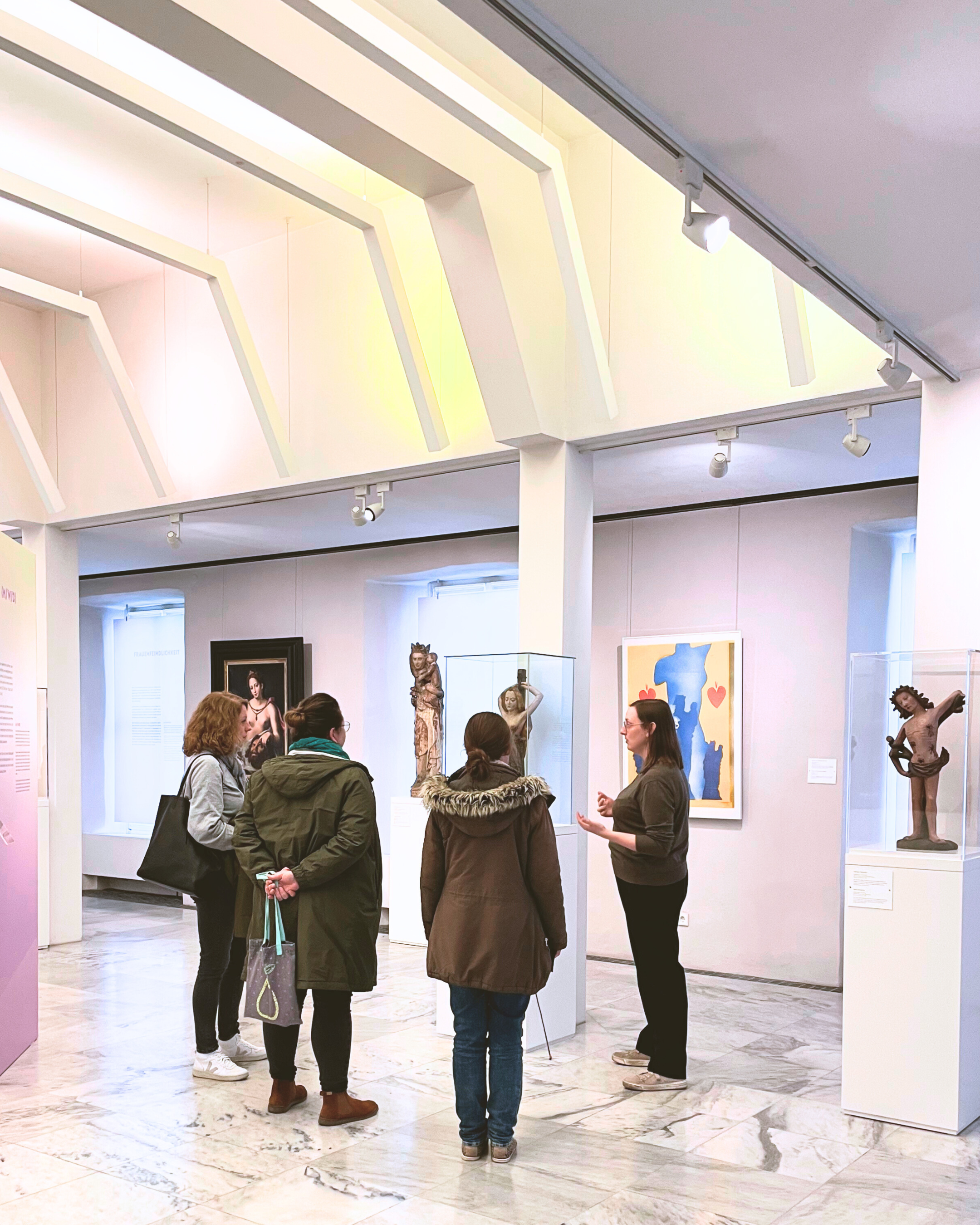 Eine vierergruppe durch die Ausstellung 'Menschenskinder' geführt. Sie stehen vor einem Kunstobjekt im Innenraum des Museums. Die Wände des Museums sind weiß, und sie wird mit verschiedenen farbigem Licht angestrahlt. Verschiedene Kunstwerke stehen im Innenraum.