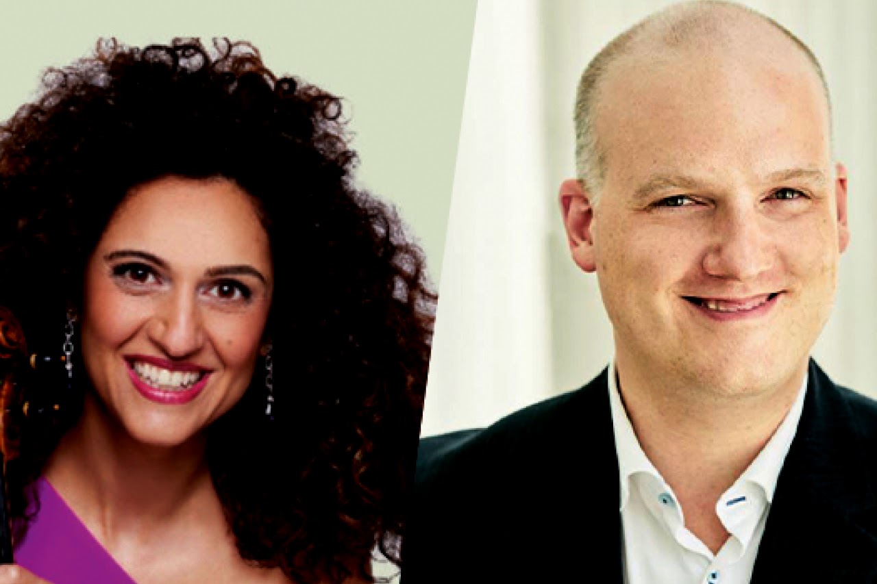 Frau mit langen dunklen Locken und Mann im schwarzen Anzug lächeln in die Kamera.