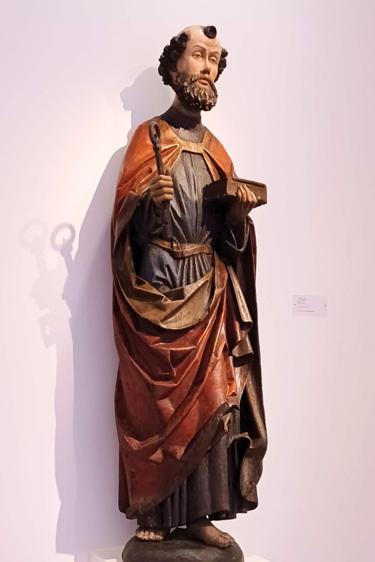 Restaurierte Petrustatue: Holzskulptur einer stehenden religiösen Figur in langer, farbig gefasster Gewandung. Die Figur hält in der rechten Hand einen Schlüssel und in der linken ein Buch. Sie steht auf einem Sockel vor einer hellen Wand.