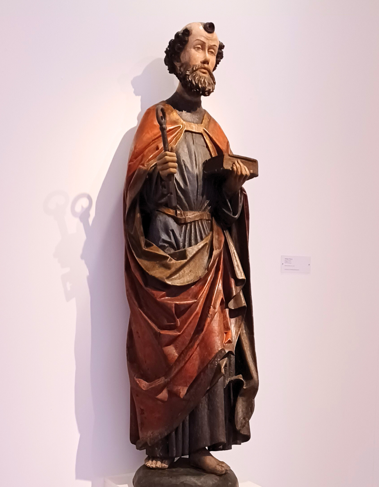 Restaurierte Petrustatue: Holzskulptur einer stehenden religiösen Figur in langer, farbig gefasster Gewandung. Die Figur hält in der rechten Hand einen Schlüssel und in der linken ein Buch. Sie steht auf einem Sockel vor einer hellen Wand.